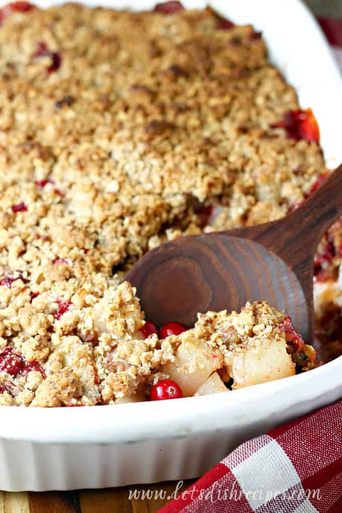 A close-up of a baked pear cranberry crisp in a white dish. A wooden spoon scoops the golden oat topping, revealing red cranberries underneath.