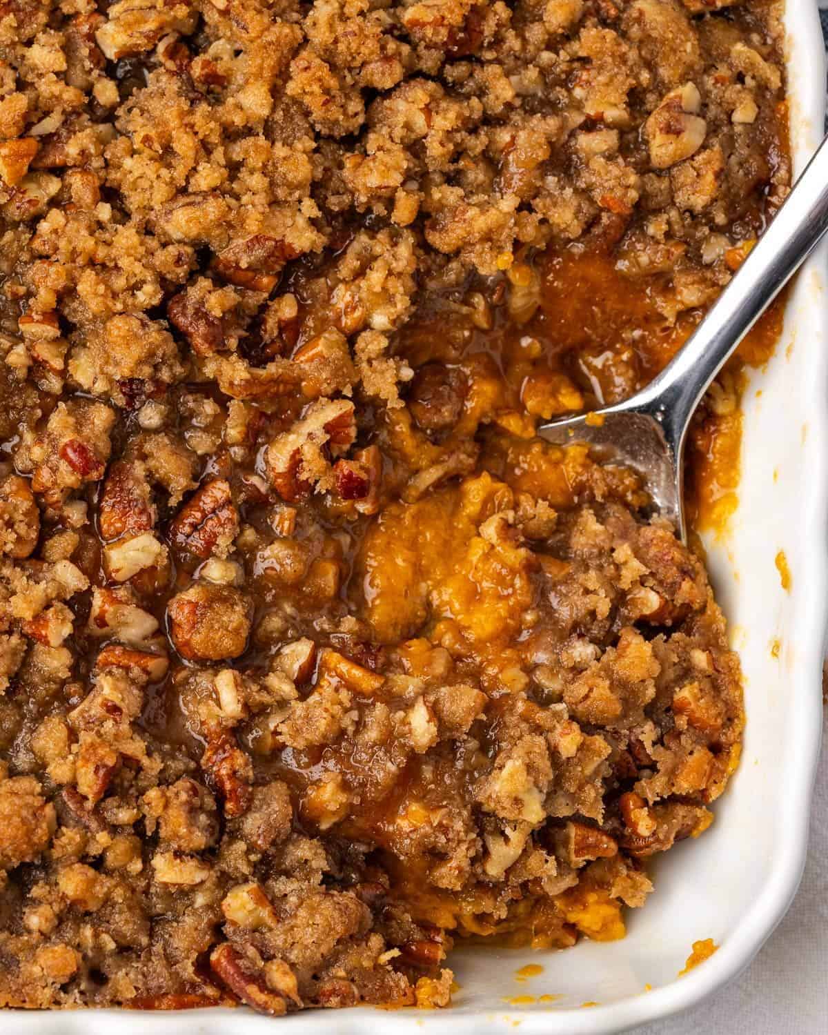 Sweet potato casserole with a crumbly pecan topping fills a white baking dish. A spoon is scooping into the rich, golden-brown mixture. Warm and inviting.