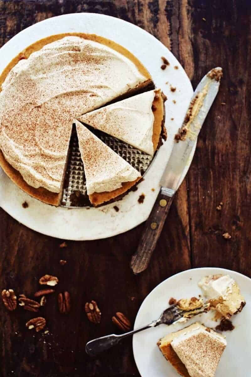 A creamy pie with a cinnamon-dusted top is partially sliced on a rustic wooden table. A slice on a white plate, surrounded by pecans, creates a cozy, inviting scene.