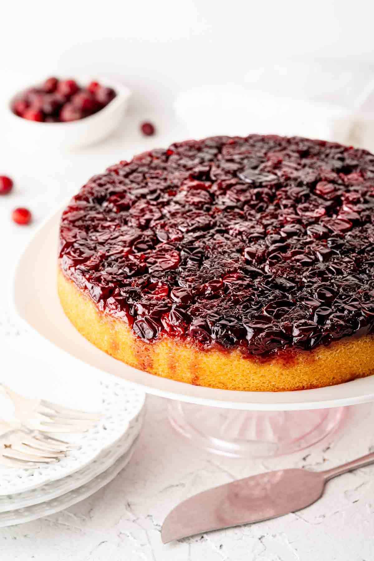 Cranberry upside-down cake on a white cake stand, displaying a glossy cranberry topping. A stack of plates, forks, and scattered cranberries are in the background.