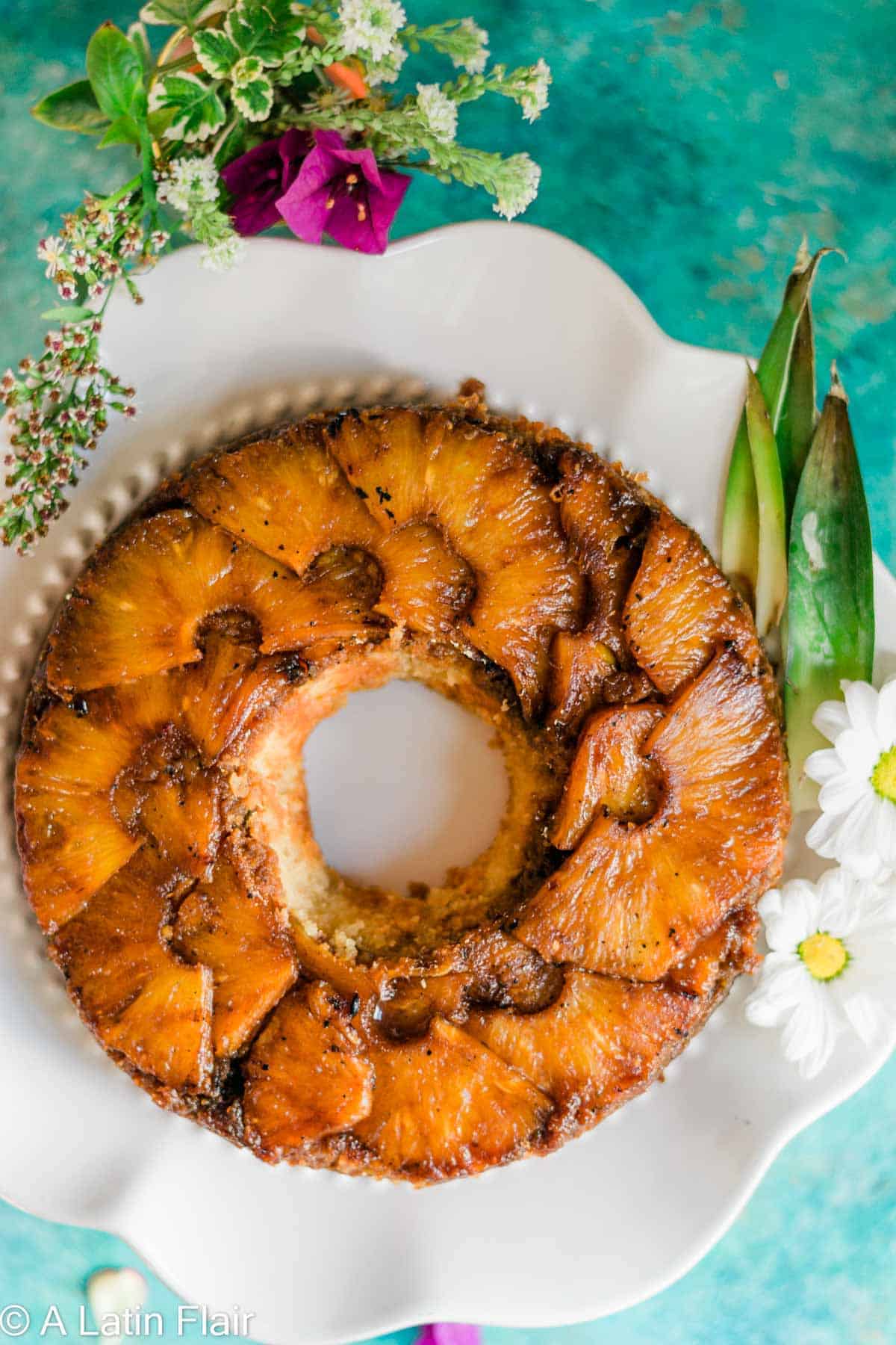 Pineapple upside-down cake on a white plate with vibrant green background, garnished with fresh flowers, creating a tropical and inviting look.