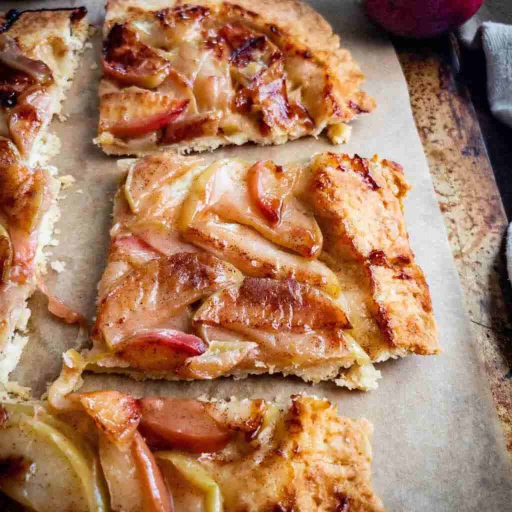 A close-up of a rustic apple tart with a flaky golden crust. The tart is topped with caramelized apple slices on parchment paper, evoking a cozy, homemade vibe.