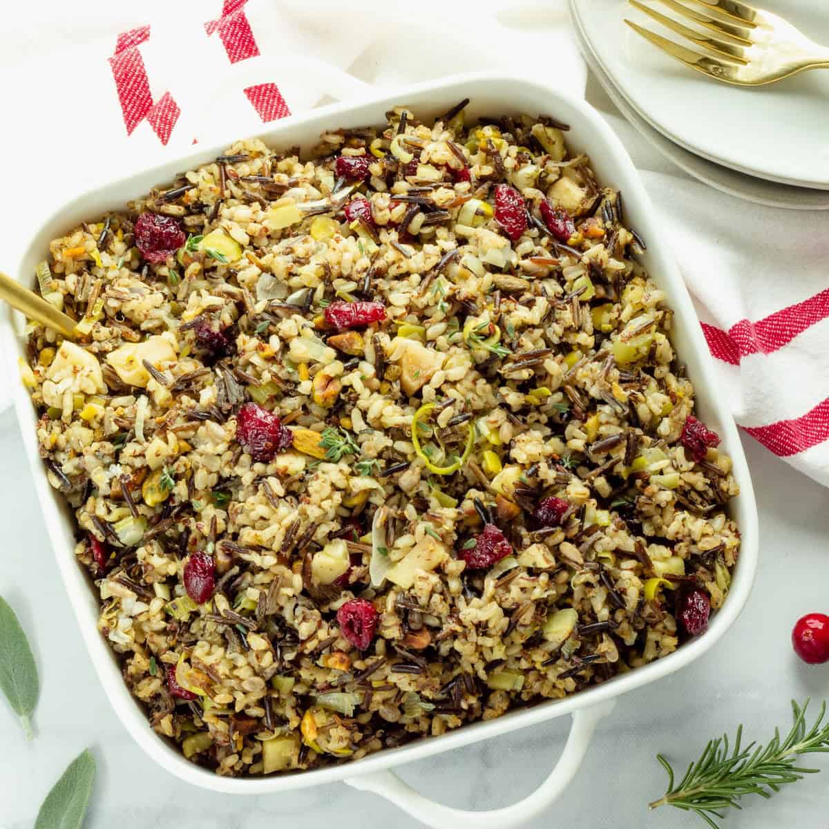 A square white dish filled with wild rice pilaf mixed with cranberries, nuts, and herbs. Nearby are plates, a fork, and a red-striped towel, creating a festive tone.