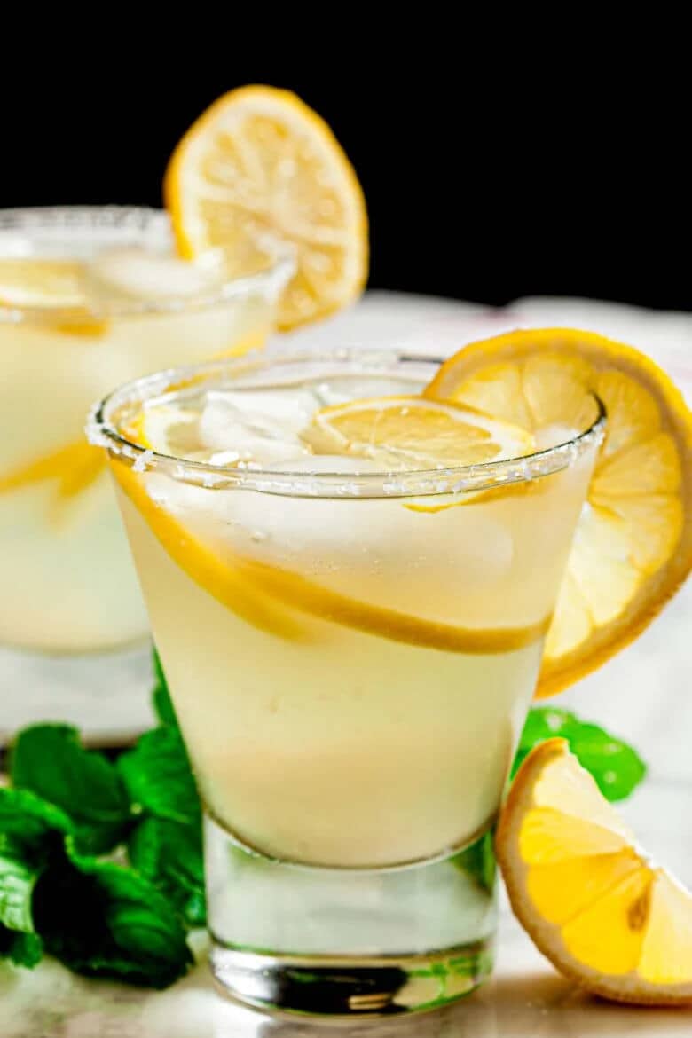 A 3-ingredient lemonade margarita in a rocks glass with a salt rim and lemon wheel garnish, with a second margarita and fresh mint in the background against a black backdrop.