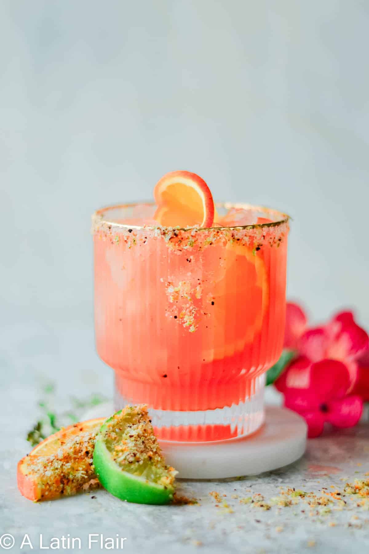 An Aperol margarita in a textured glass with a colorful spiced sugar rim, garnished with a grapefruit slice, alongside lime and grapefruit wedges coated in Tajín seasoning and pink flowers.