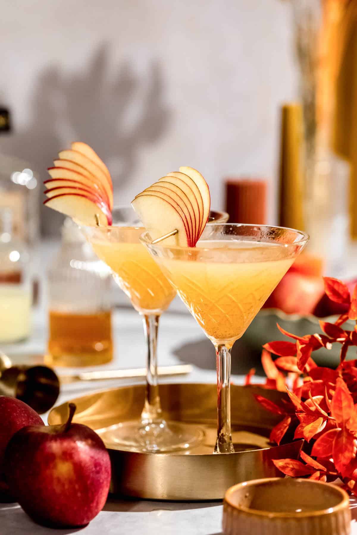 Two golden apple cider martinis in martini glasses garnished with decorative fan-shaped apple slices, displayed on a gold tray with fresh red apples and autumn leaves.