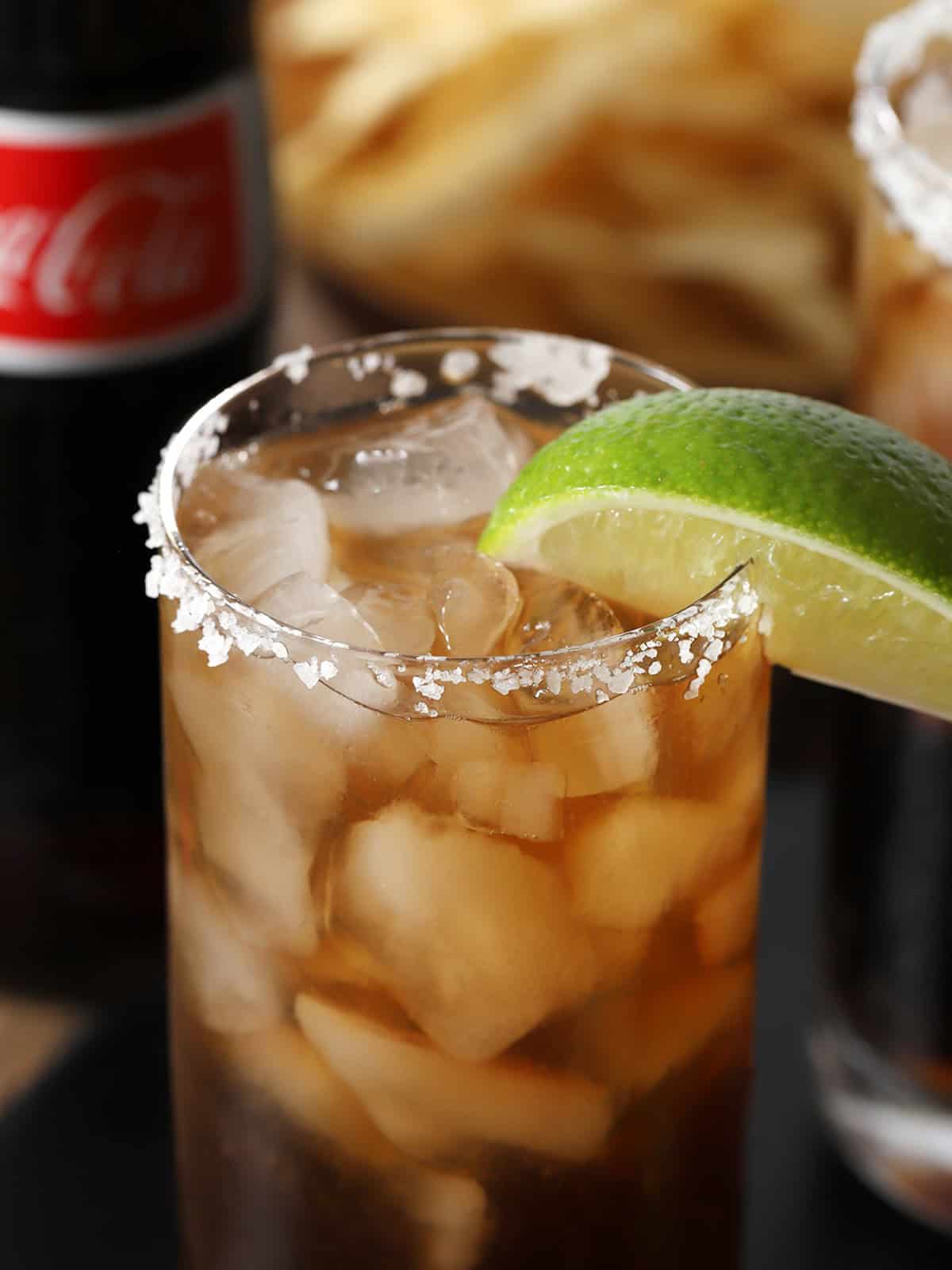 A Batanga cocktail in a rocks glass with a salt rim and lime wedge garnish, filled with ice and showing the golden-brown color of tequila mixed with cola, with a Coca-Cola bottle visible in the background.