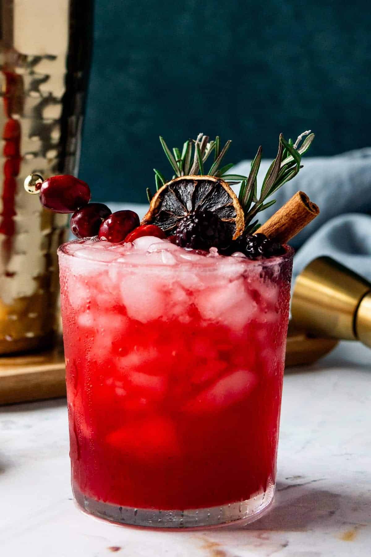 Vibrant red blackberry smash cocktail in a rocks glass filled with crushed ice, garnished with skewered fresh cranberries, dried citrus wheel, fresh rosemary, cinnamon stick, and blackberry, with a gold cocktail shaker in the background.