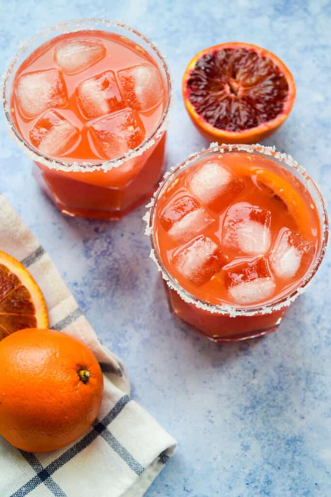 Two blood orange margaritas in rocks glasses with salt rims and large ice cubes, surrounded by fresh whole and halved blood oranges on a blue surface with a striped kitchen towel.