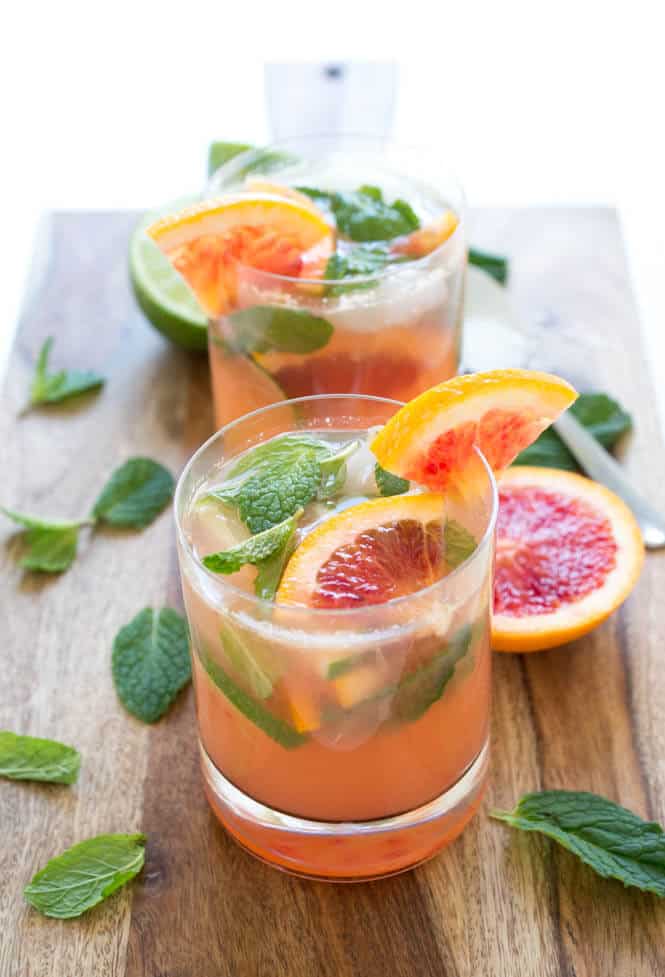 Two glasses of blood orange mojitos with coral-pink liquid filled with ice, muddled fresh mint leaves, and lime, garnished with blood orange slices and mint sprigs, displayed on a rustic wooden board with scattered fresh mint leaves, blood orange halves, and a lime wedge.