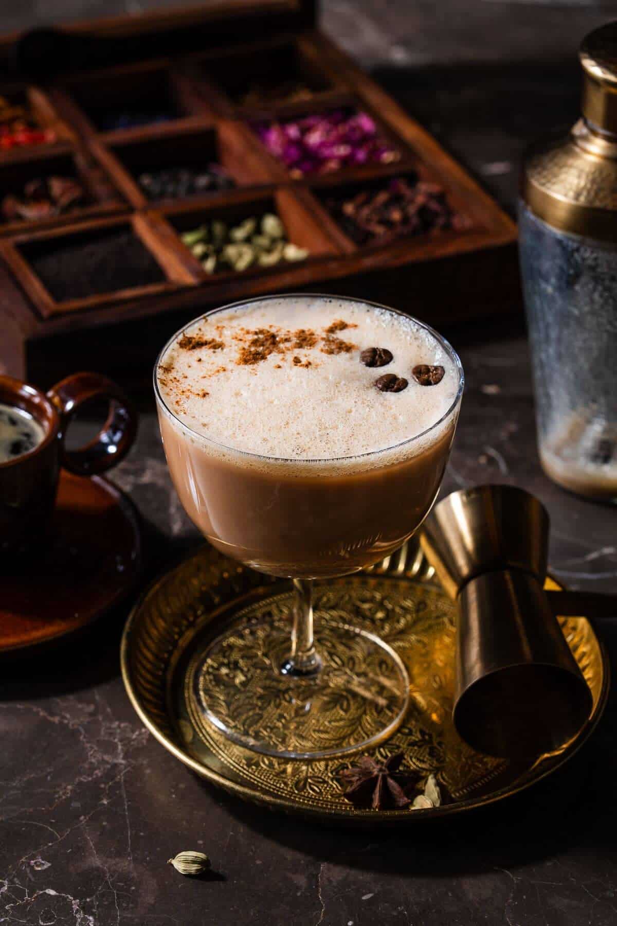 Layered chai espresso martini in a coupe glass with frothy top dusted with cinnamon and garnished with three coffee beans, served on an ornate gold tray with cocktail jigger, spoon, and star anise, with a wooden spice box containing chai spices in the background.