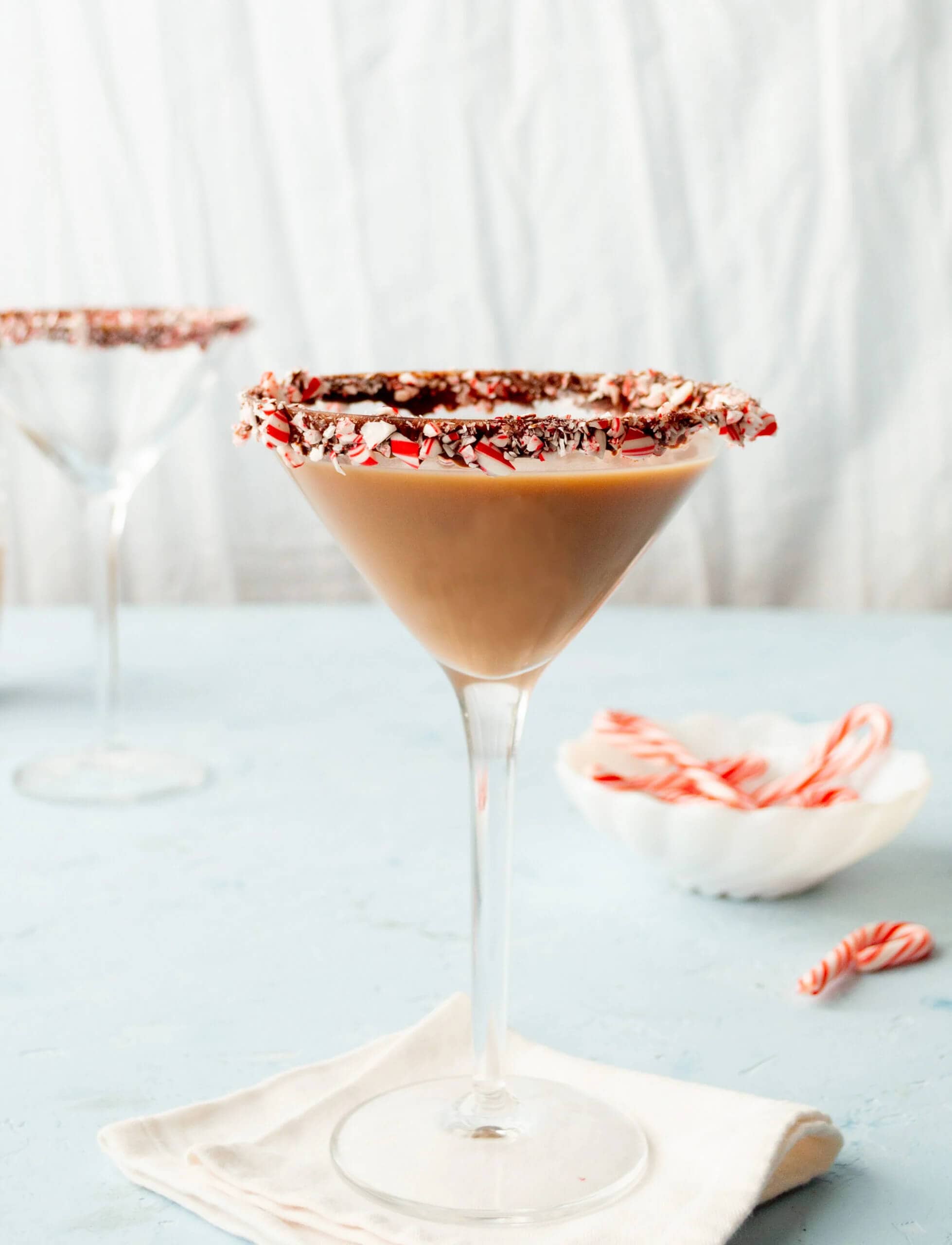Creamy chocolate peppermint martini in a martini glass with a rim coated in crushed candy cane and chocolate, with additional candy canes on a white plate in the background.