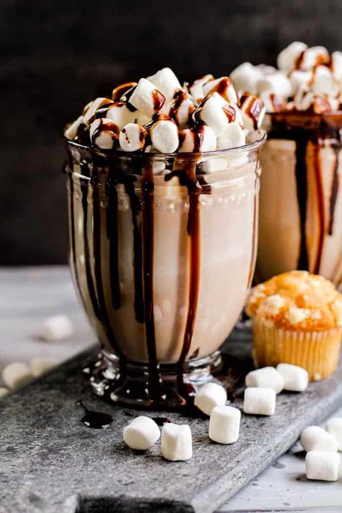 Glass of chocolate coquito with creamy light brown liquid in a ridged mug, topped with mini marshmallows and drizzled with chocolate sauce running down the sides, served on a gray slate surface with scattered marshmallows and a muffin against a dark background.