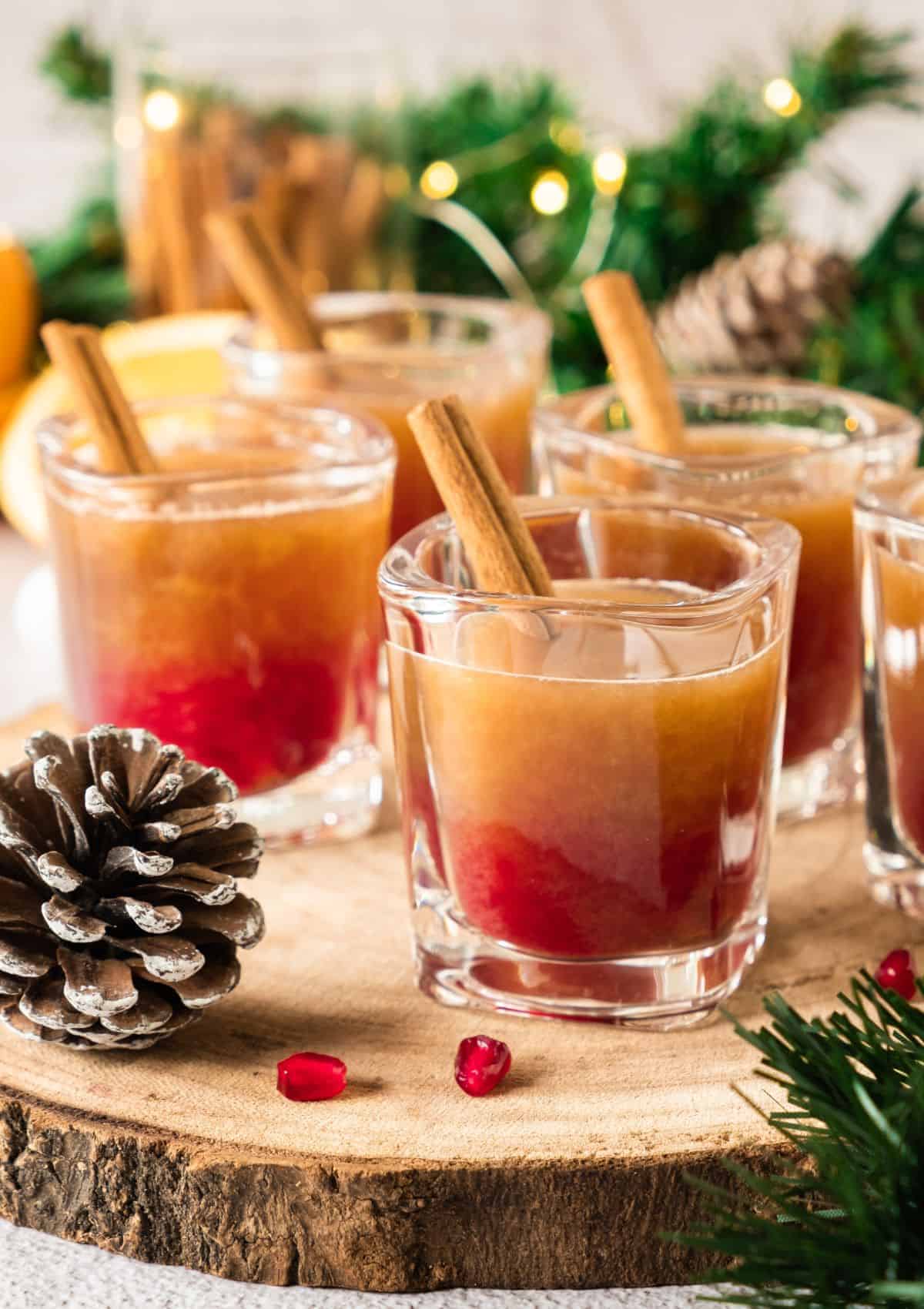 Layered Christmas ginger syrup cocktails in rocks glasses with red and golden amber layers, garnished with cinnamon sticks, displayed on a wood slice with pine cones, pomegranate arils, evergreen branches, and twinkling lights in the background.