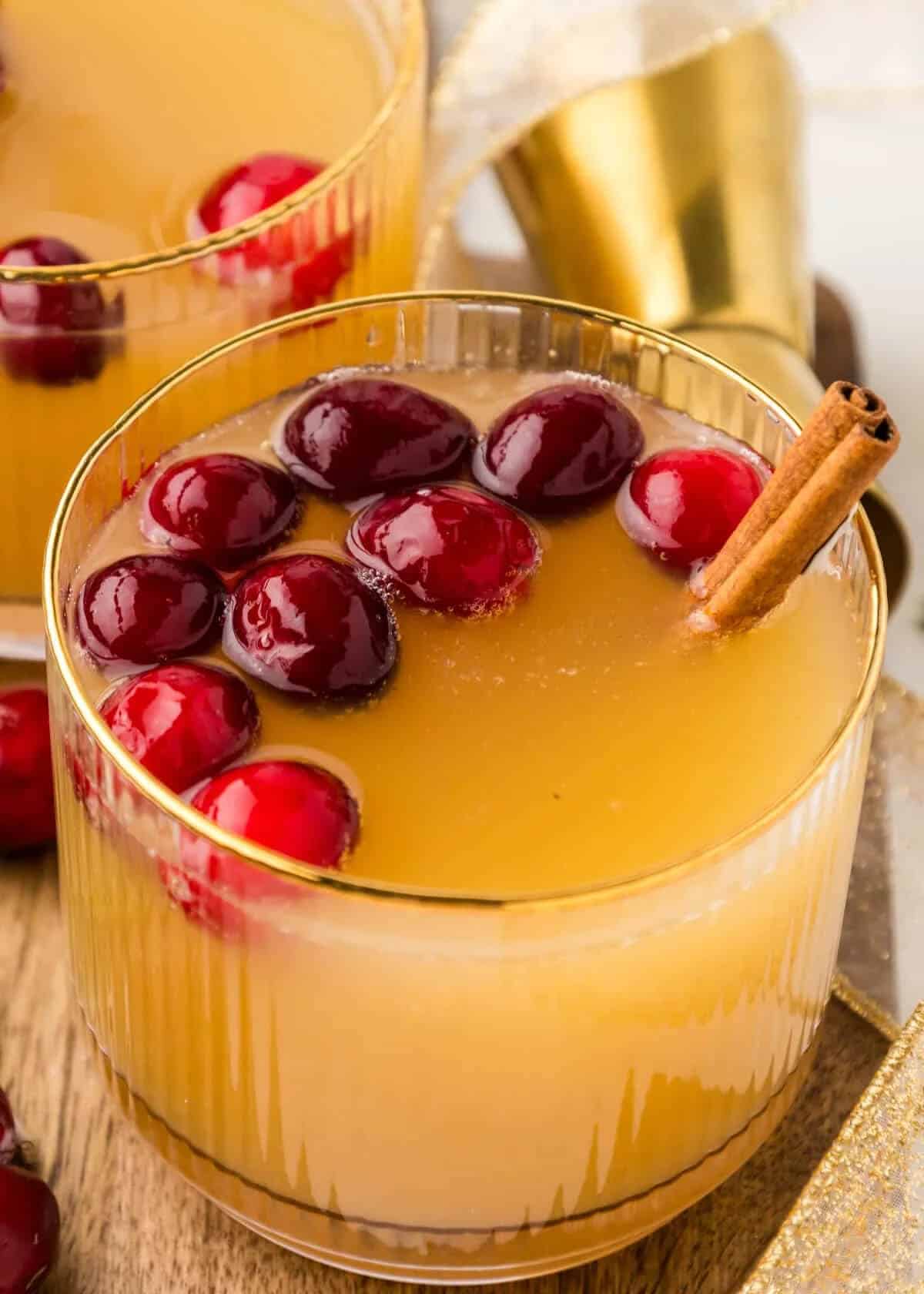 Christmas old fashioned cocktail in a gold-rimmed glass, garnished with fresh cranberries arranged in a wreath pattern and a cinnamon stick, with additional cocktails and a gold shaker in the background.