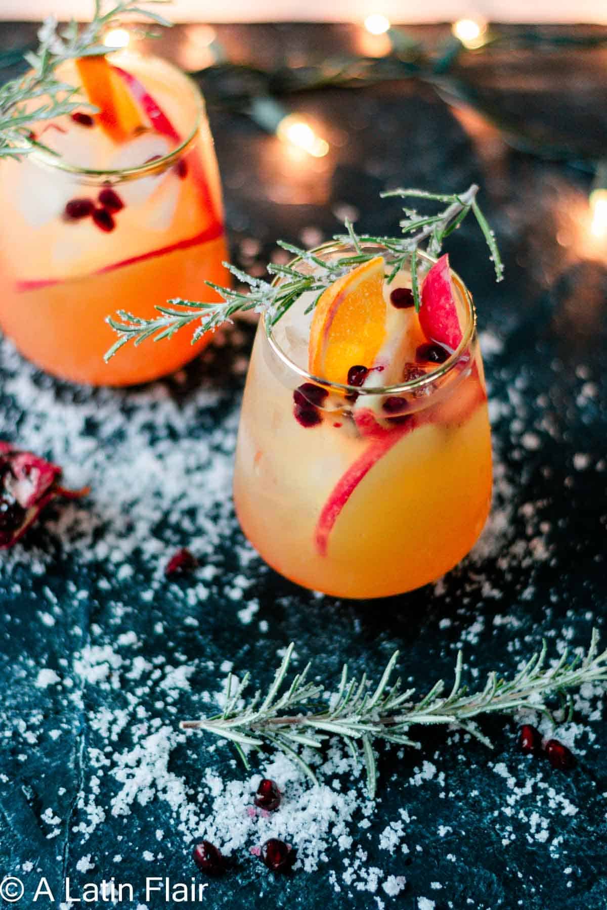 Two festive glasses of citrusy holiday sangria filled with ice, orange slices, apple slices, and pomegranate seeds. Each glass is garnished with a frosted rosemary sprig. The dark surface beneath is sprinkled with faux snow, with warm string lights glowing softly in the background.