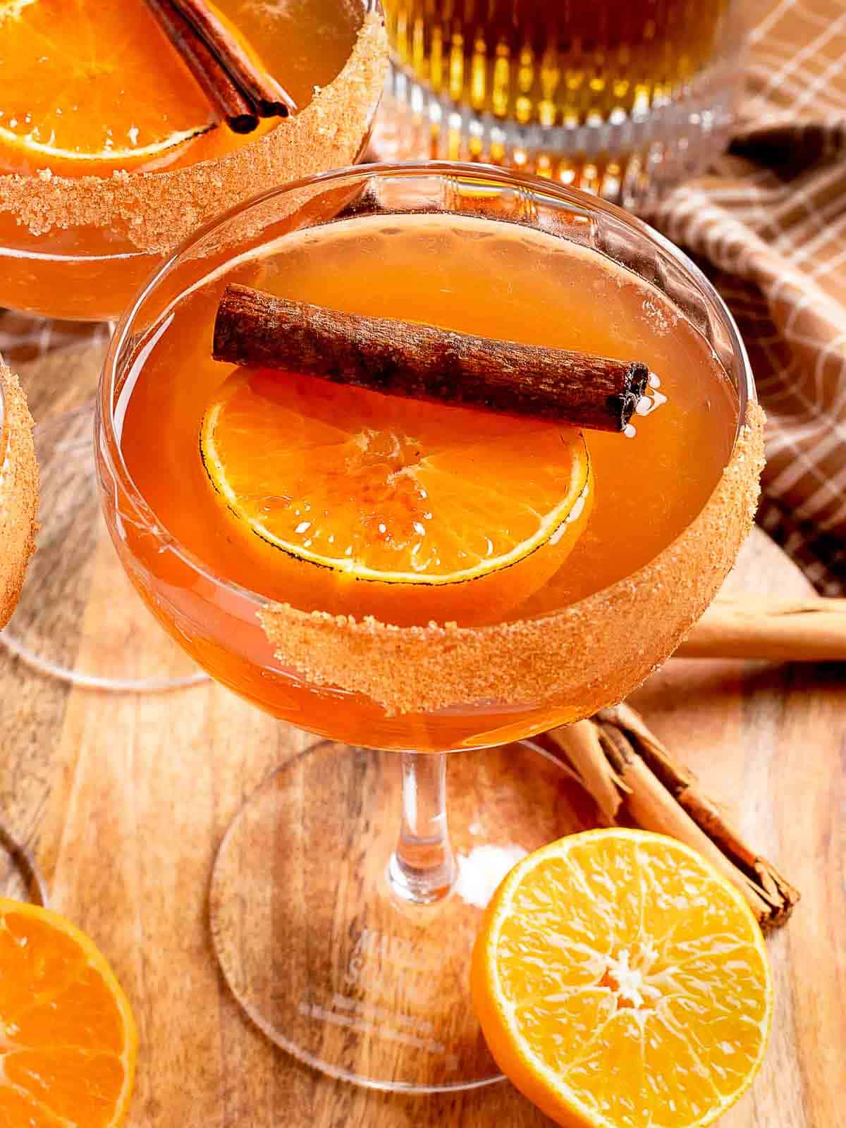 Amber-colored cinnamon old fashioned cocktail in a coupe glass with cinnamon-sugar rim, garnished with an orange slice and cinnamon stick, surrounded by fresh orange slices and cinnamon sticks on a wooden surface.