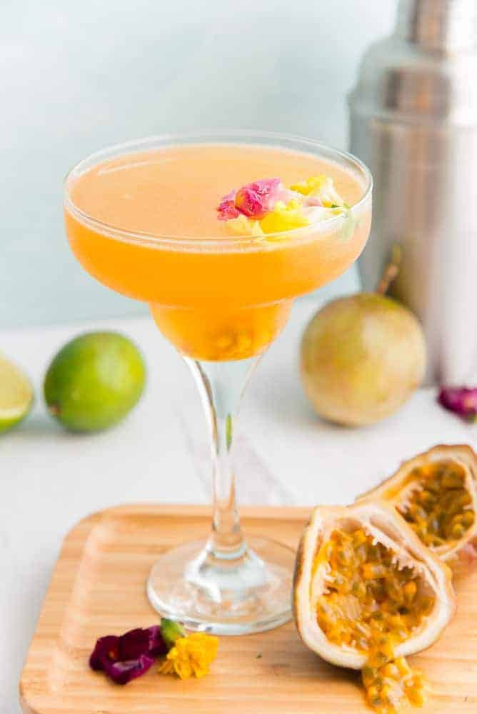 Classic passion fruit daiquiri in a martini glass with golden orange liquid, garnished with pink and yellow edible flowers, served on a wooden board alongside fresh passion fruit halves, with limes and a cocktail shaker in the background.