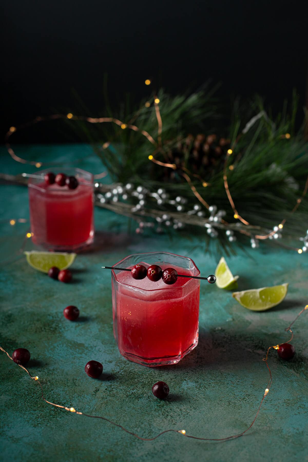 Two cranberry gin and tonics in rocks glasses garnished with skewered fresh cranberries, surrounded by lime wedges, evergreen branches with pinecone, silver beaded garland, and twinkling string lights on a teal surface.