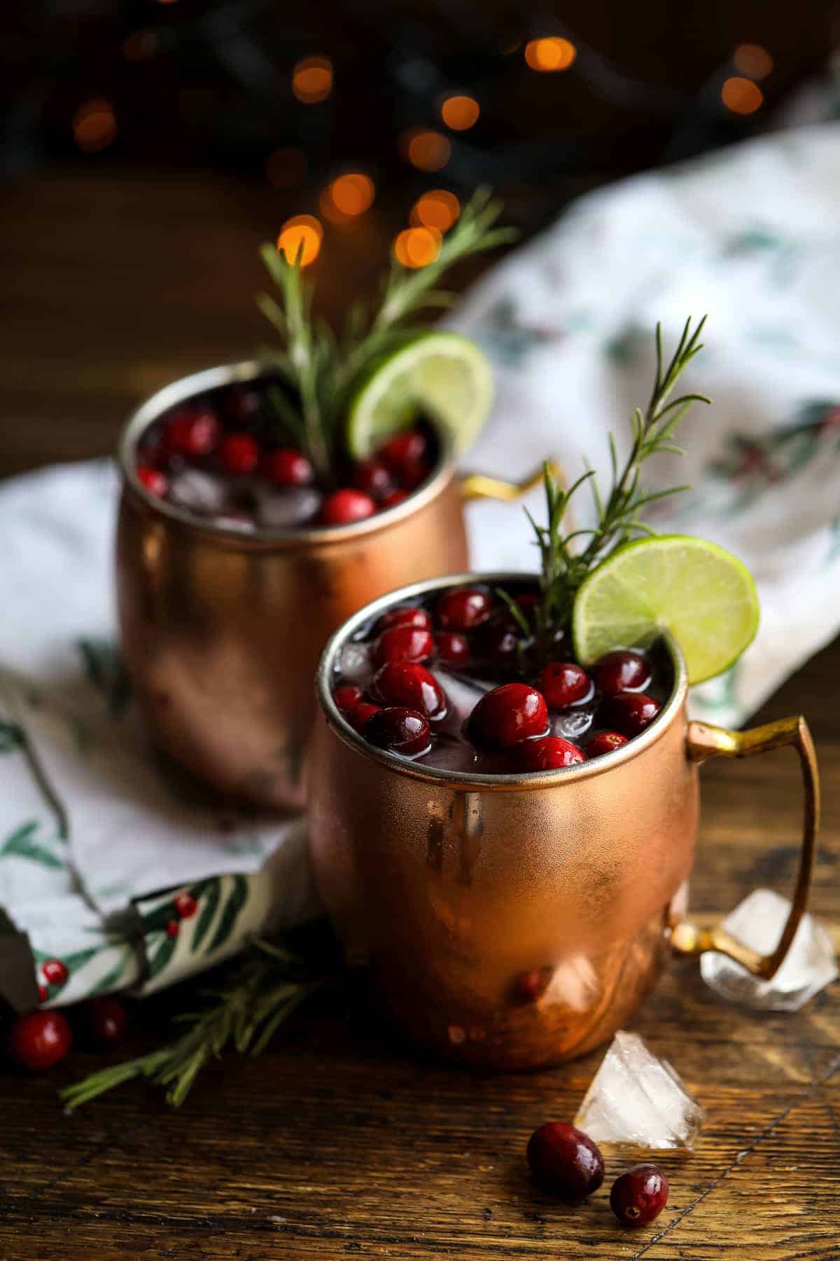 Cranberry Moscow mules in copper mugs filled with ice and fresh cranberries, garnished with lime wedges and fresh rosemary sprigs, displayed with evergreen branches and warm bokeh lights in the background.