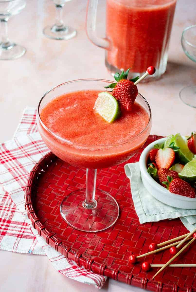 Frozen strawberry rum-rita (rum margarita) in a margarita glass garnished with a lime wedge and fresh strawberry, served on a red woven tray with a checkered napkin, alongside a blender of the cocktail mixture and a bowl of fresh strawberries and lime wedges.