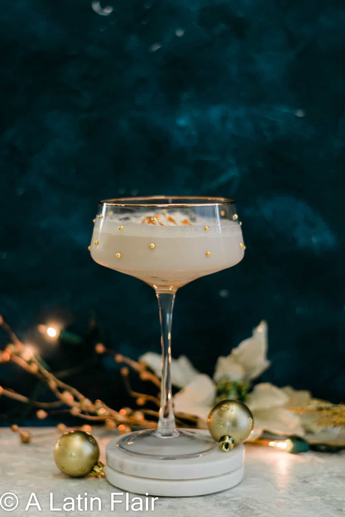 Creamy dulce de leche martini in a gold-rimmed coupe glass decorated with gold pearl dragees and caramel drizzle, displayed on a white coaster with festive holiday decorations including gold ornaments, white poinsettias, and berry branches against a dark teal background.