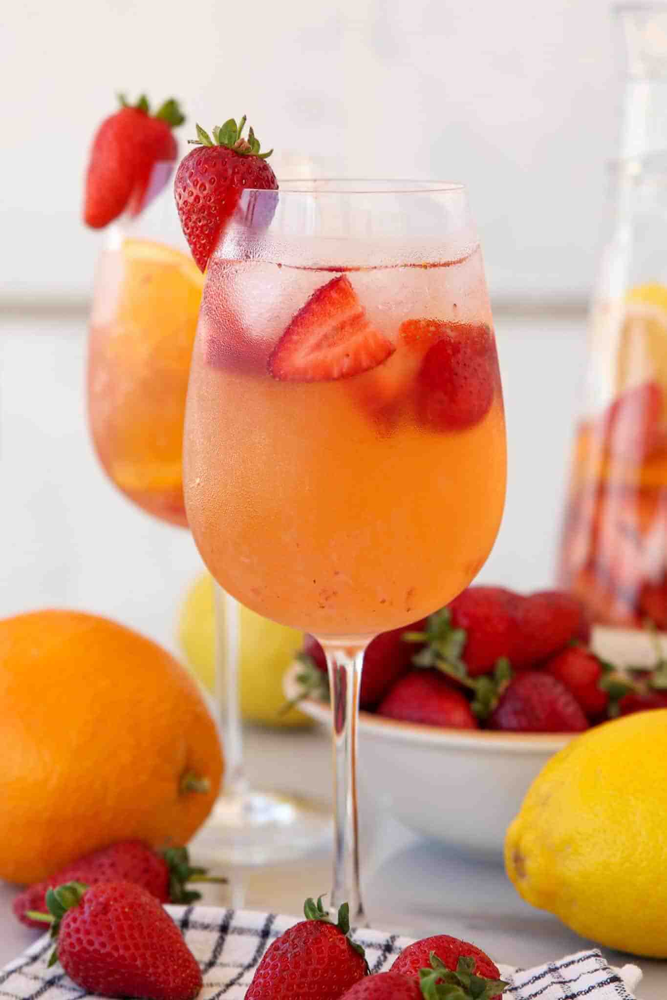 Glass of easy strawberry sangria with peachy-orange liquid filled with ice and fresh strawberry slices, garnished with a whole strawberry on the rim, displayed on a white surface with fresh strawberries, oranges, and lemons scattered around, and additional sangria glasses in the background.