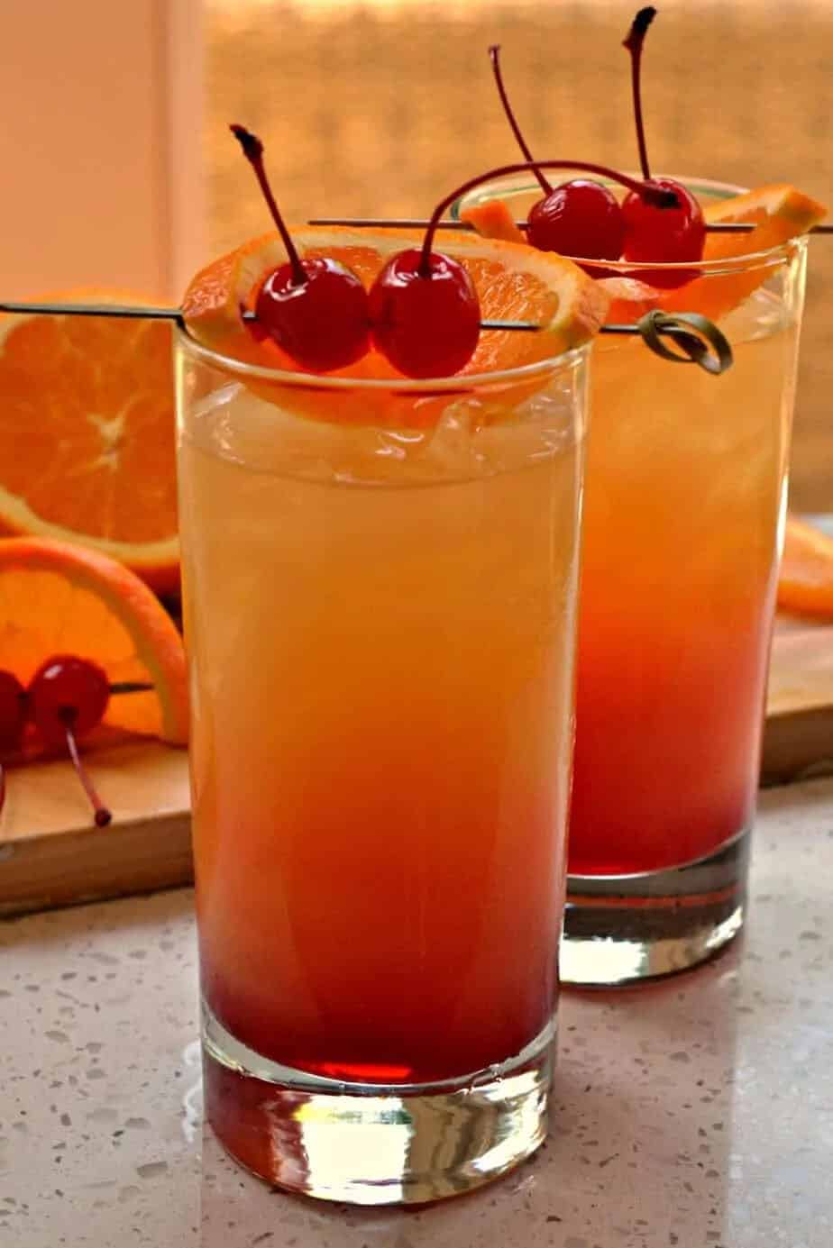 Easy tequila sunrise cocktails in tall glasses with the signature red to orange gradient, garnished with orange slices and maraschino cherries, with fresh orange slices and cherries on a wooden board in the background.