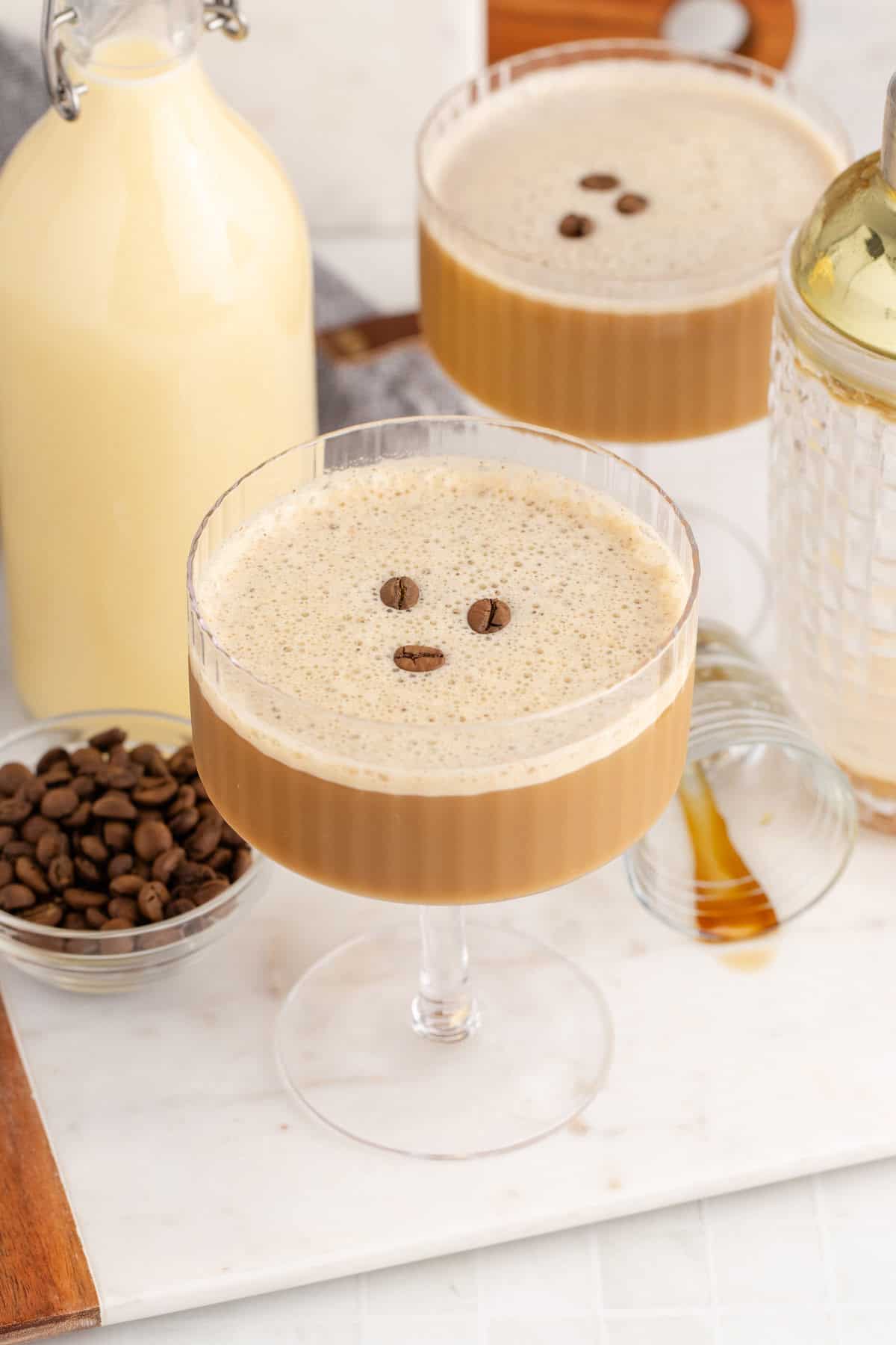 Two layered eggnog espresso martinis in coupe glasses with golden coffee layer and white frothy top, each garnished with three coffee beans, displayed with a bottle of eggnog and bowl of coffee beans.