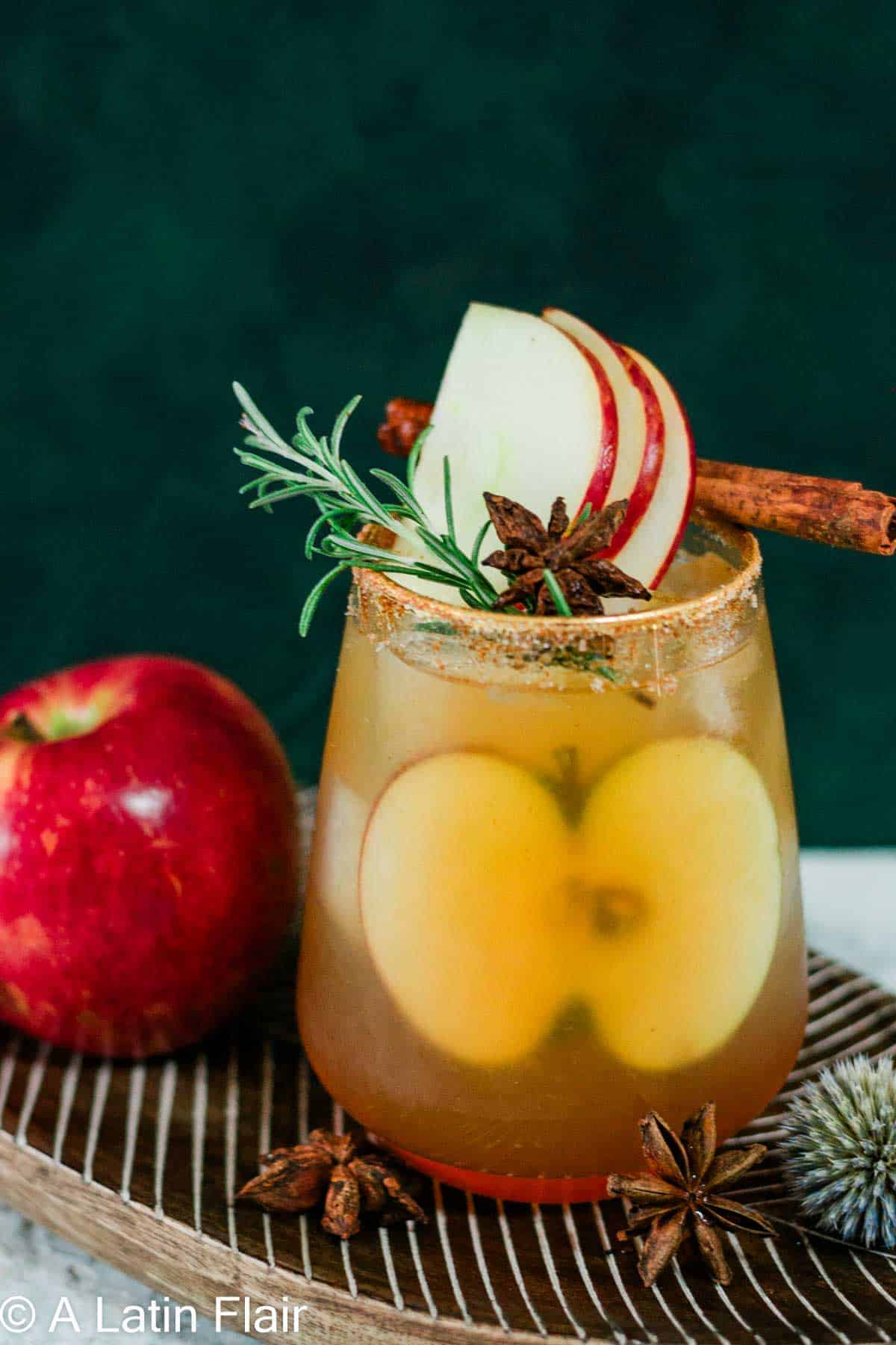 A spiced apple margarita is served in a short glass with a cinnamon-sugar rim of the glass. Thin apple slices are visible inside the drink, while the top is garnished with a fresh apple slice, a sprig of rosemary, star anise, and a cinnamon stick. The cocktail sits on a wooden tray surrounded by whole apples and warm spices, set against a dark moody background for a cozy fall-inspired look.