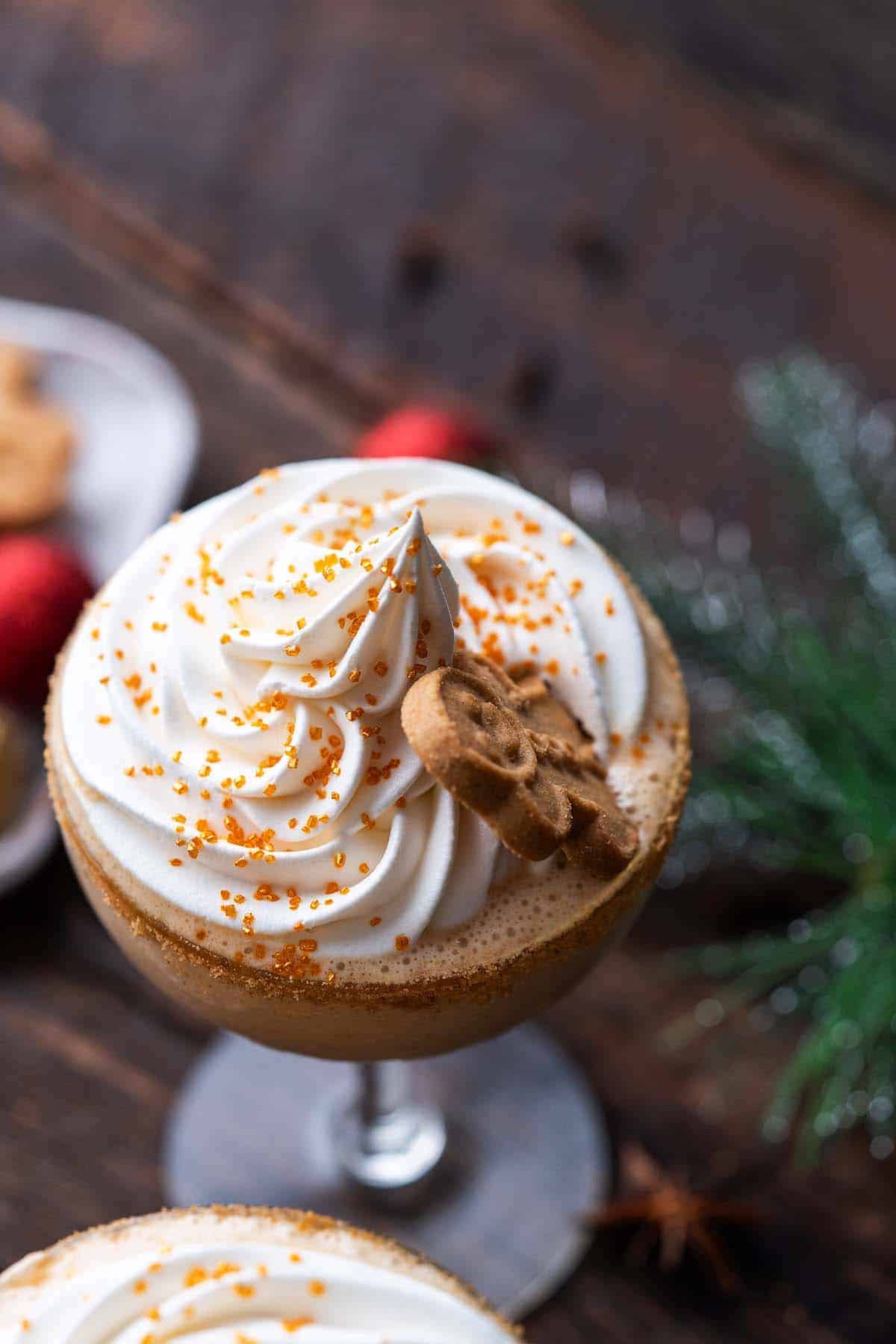 Creamy gingerbread cocktail topped with swirled whipped cream, gold sugar sprinkles, and garnished with a gingerbread man cookie and silver Christmas tree decoration, with holiday ornaments and evergreen in the background.