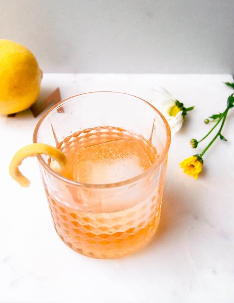A grapefruit-infused tequila old fashioned in a textured rocks glass with a large ice sphere and a grapefruit peel twist, with fresh grapefruit and yellow flowers on a white marble surface.