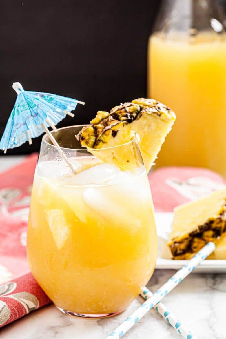 Glass of Hawaiian Mai Tai with ice and golden tropical liquid, garnished with a fresh pineapple wedge and blue paper cocktail umbrella with blue striped straws, displayed on a white marble surface with a coral patterned napkin, pitcher of Mai Tai mix and pineapple slices in the background.