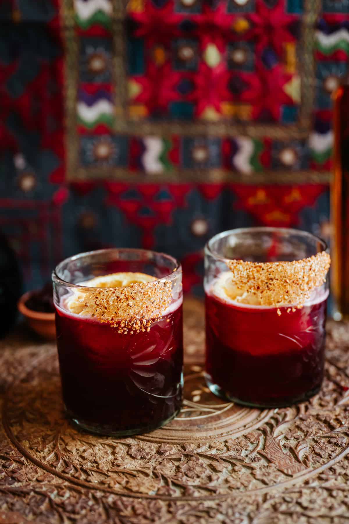 Hibiscus margarita recipe (Jamaica margarita) with golden sugar rim in rocks glasses, served on carved wooden tray with colorful textile backdrop.