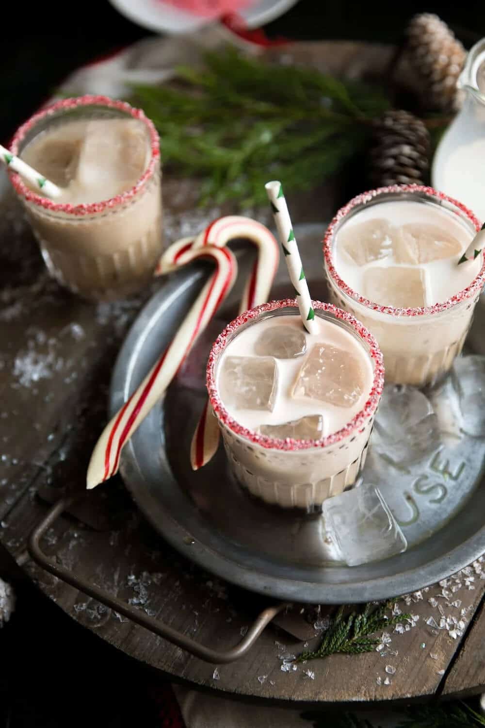 Creamy peppermint white Russian cocktails in rocks glasses with crushed candy cane rims and large ice cubes, served with striped green and white paper straws on a dark tray surrounded by candy canes, pine cones, and evergreen branches.