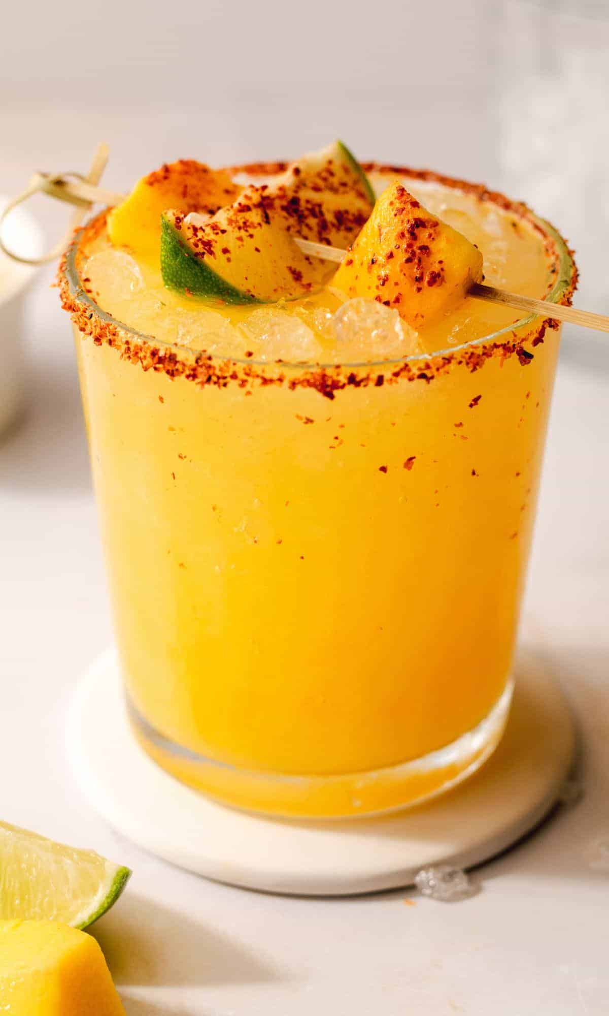 A hot honey spicy mango margarita in a rocks glass with a chili lime salt rim, garnished with fresh mango chunks and lime wedges sprinkled with chili flakes, served on a white marble coaster.