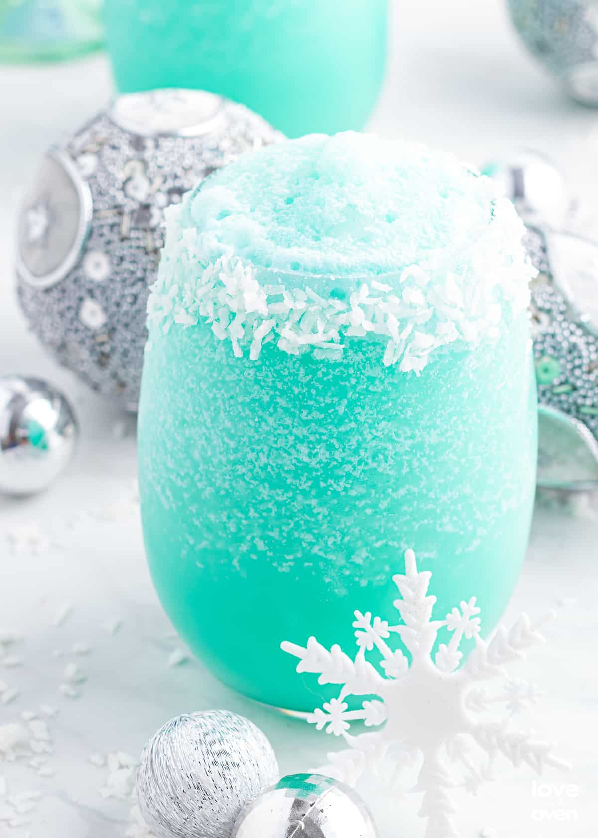 Bright turquoise Jack Frost frozen cocktail in a stemless wine glass with a coconut-rimmed edge and white snowflake decoration, surrounded by silver holiday ornaments and additional cocktails in the background.