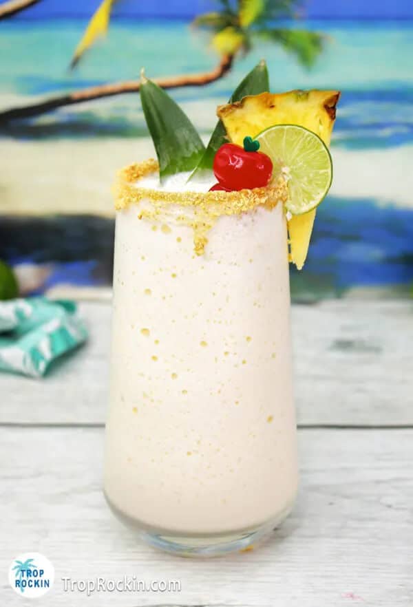 Frozen key lime colada in a tall glass with a graham cracker rim, garnished with a pineapple wedge, lime wheel, pineapple leaf, and maraschino cherry, displayed on a white wooden surface with a tropical beach scene backdrop and a small teal gift box.