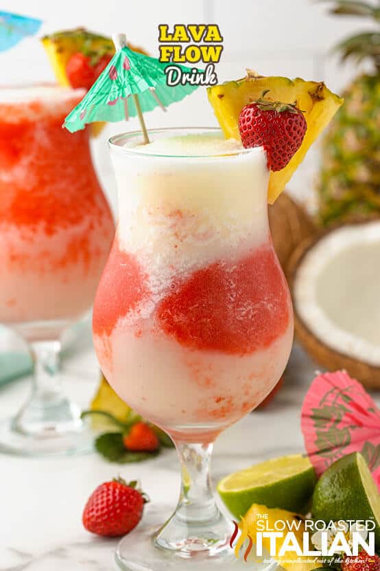 Frozen lava flow cocktail in a hurricane glass with layered white coconut piña colada and red strawberry swirls creating a marbled effect, garnished with a pineapple wedge, fresh strawberry, and teal paper umbrella, displayed with fresh pineapple, coconut, limes, and strawberries on a white surface.