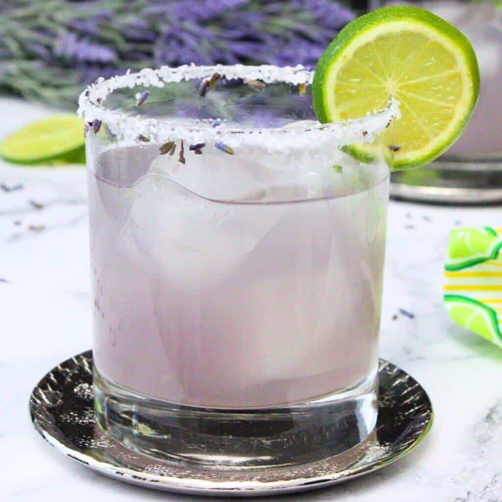 Lavender margarita recipe with sugar and dried lavender rim, garnished with fresh lime wheel on silver coaster with lavender sprigs.