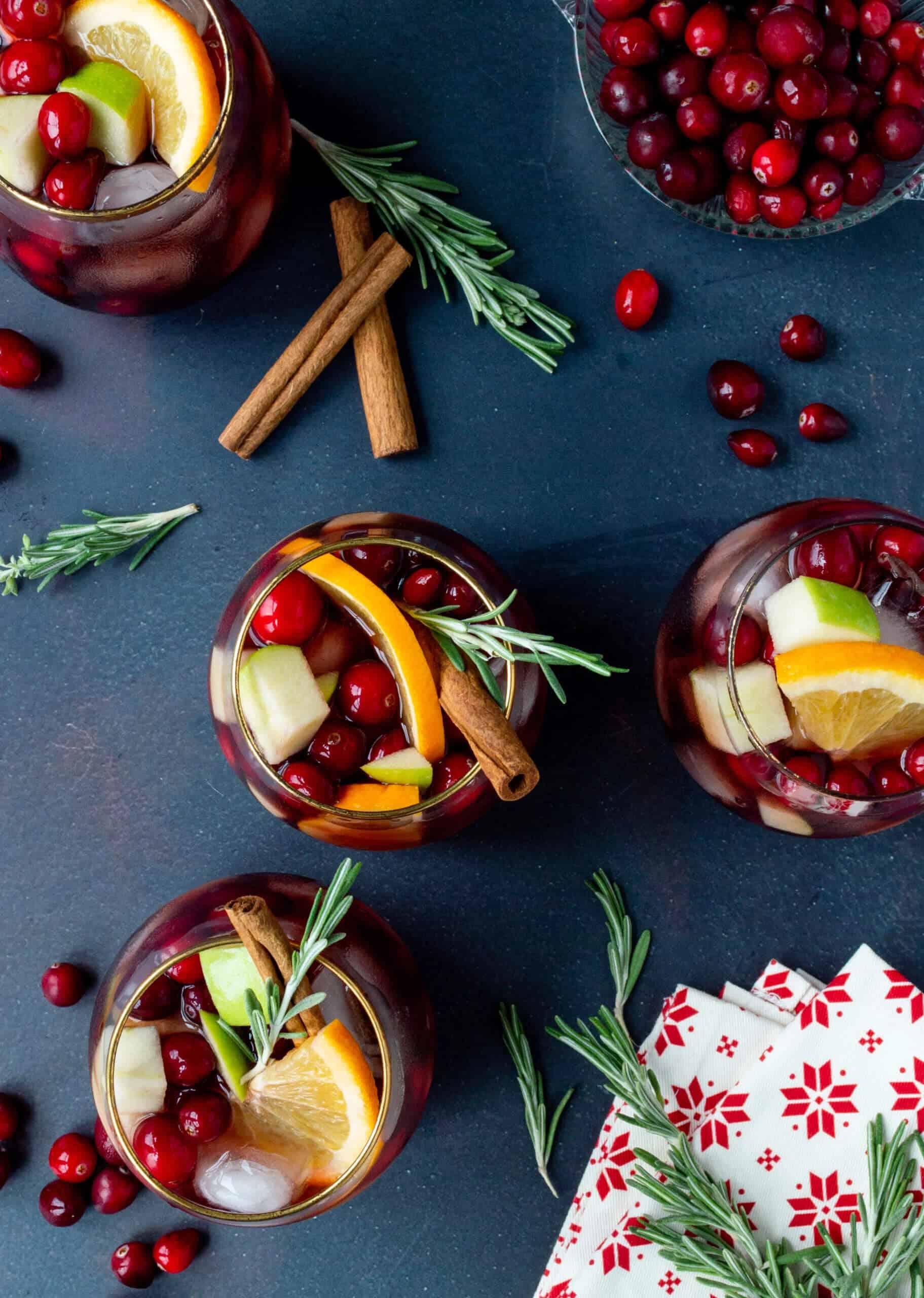 Overhead view of red sangria glasses garnished with orange slices, apple chunks, fresh cranberries, cinnamon sticks, and rosemary sprigs, arranged on a dark blue surface with a bowl of cranberries and holiday napkin.