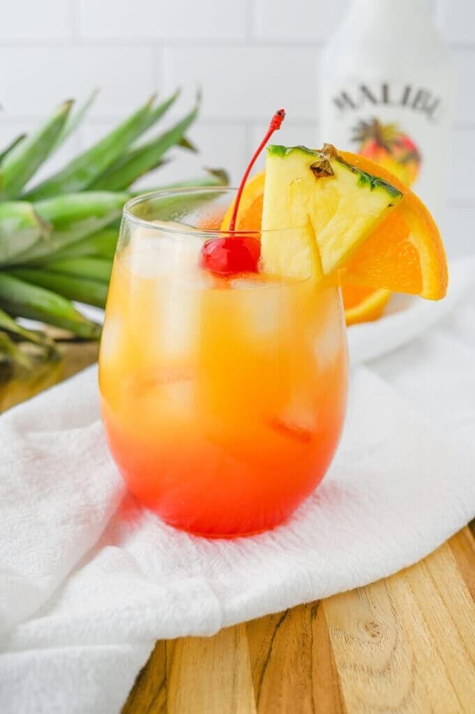 Malibu Sunset cocktail in a stemless wine glass with a vibrant layered sunset gradient from red to orange to yellow, filled with ice and garnished with a pineapple wedge, orange slice, and maraschino cherry with a red straw, displayed on a white cloth with tropical palm leaves and a Malibu rum bottle in the background.