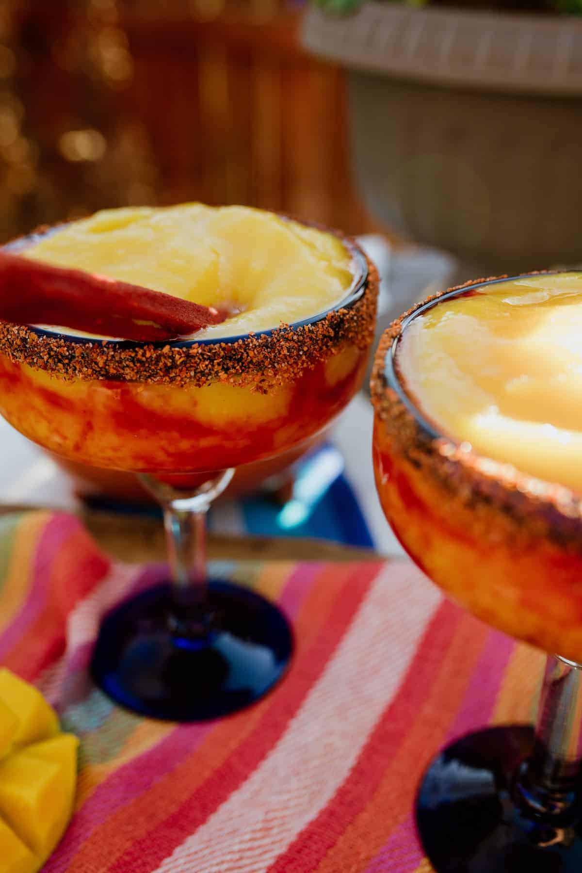Mangonada margarita recipe with chamoy and Tajín rim in blue-stemmed glasses, layered with mango and chamoy swirls.