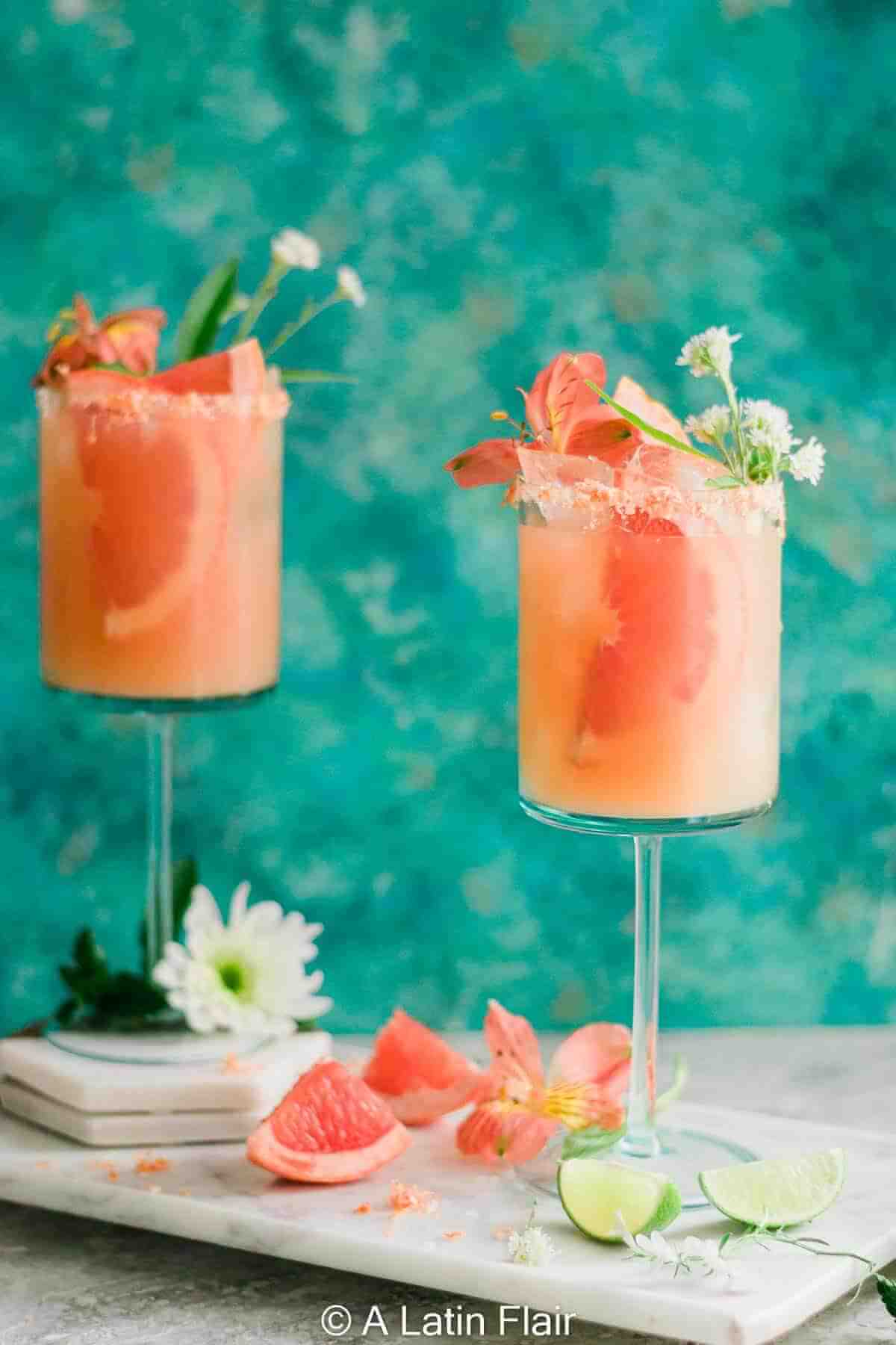 Two elegant pink citrus cocktails are served in tall stemmed glasses with coarse salt on the rim of the glass. Each drink is garnished with fresh grapefruit slices and delicate white flowers. The cocktails sit on a light marble board with sliced grapefruit and lime wedges scattered around. The background is a textured turquoise wall, giving the scene a bright, fresh, and summery feel.