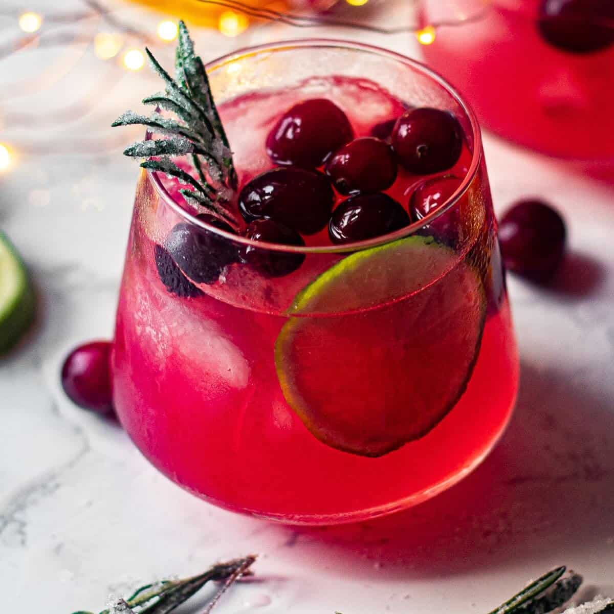 Mistletoe margarita recipe with fresh cranberries, lime wheel, and sugared rosemary garnish in rocks glass with festive holiday lights.