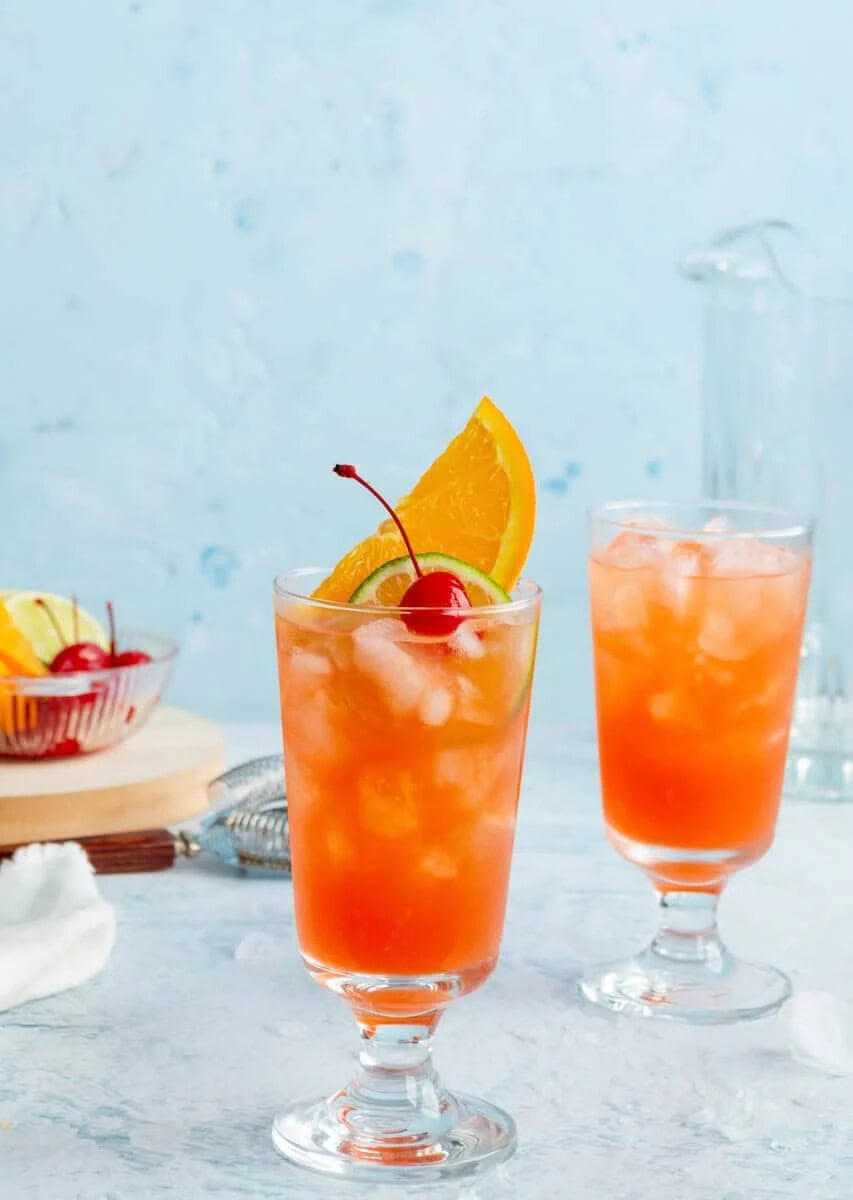 Two glasses of New Orleans Hurricane cocktails in footed glasses filled with ice and bright orange-red liquid, garnished with orange slices and maraschino cherries, displayed on a white marble surface with a light blue background and a small bowl of fresh fruit garnishes.