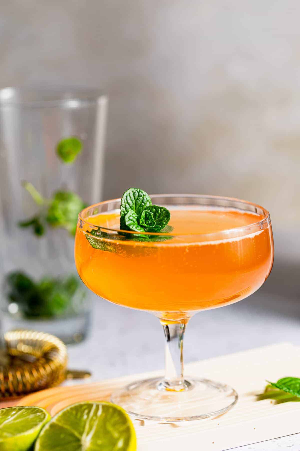 Old Cuban cocktail in a coupe glass with a golden amber color, garnished with fresh mint leaves and a lime wheel, displayed on a white surface with sliced limes, a honey dipper, and fresh mint leaves in the background.
