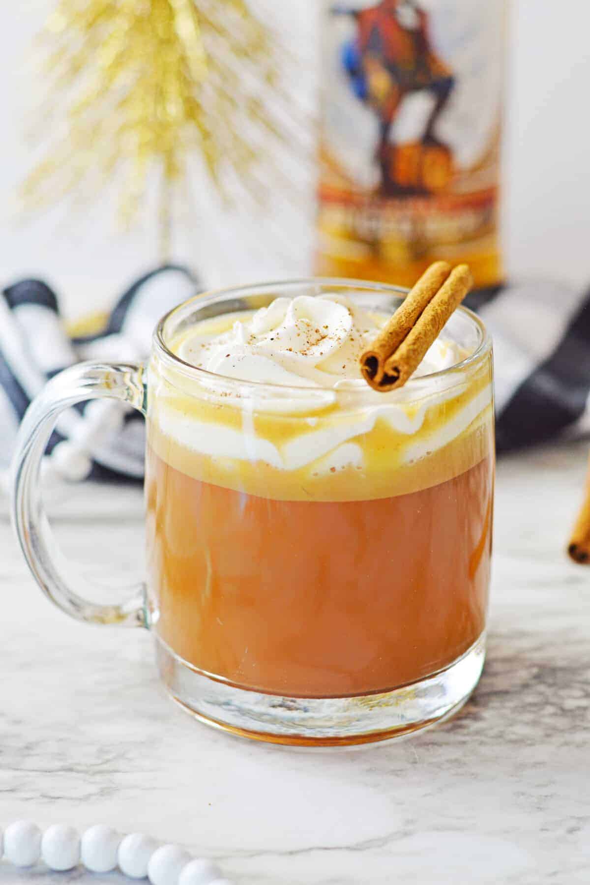 Glass mug of old-fashioned hot buttered rum with layered amber liquid, melted butter, and whipped cream topping, garnished with two cinnamon sticks and dusted with nutmeg, displayed on a white marble surface with a rum bottle, buffalo plaid fabric, and white pom pom garland in the background.