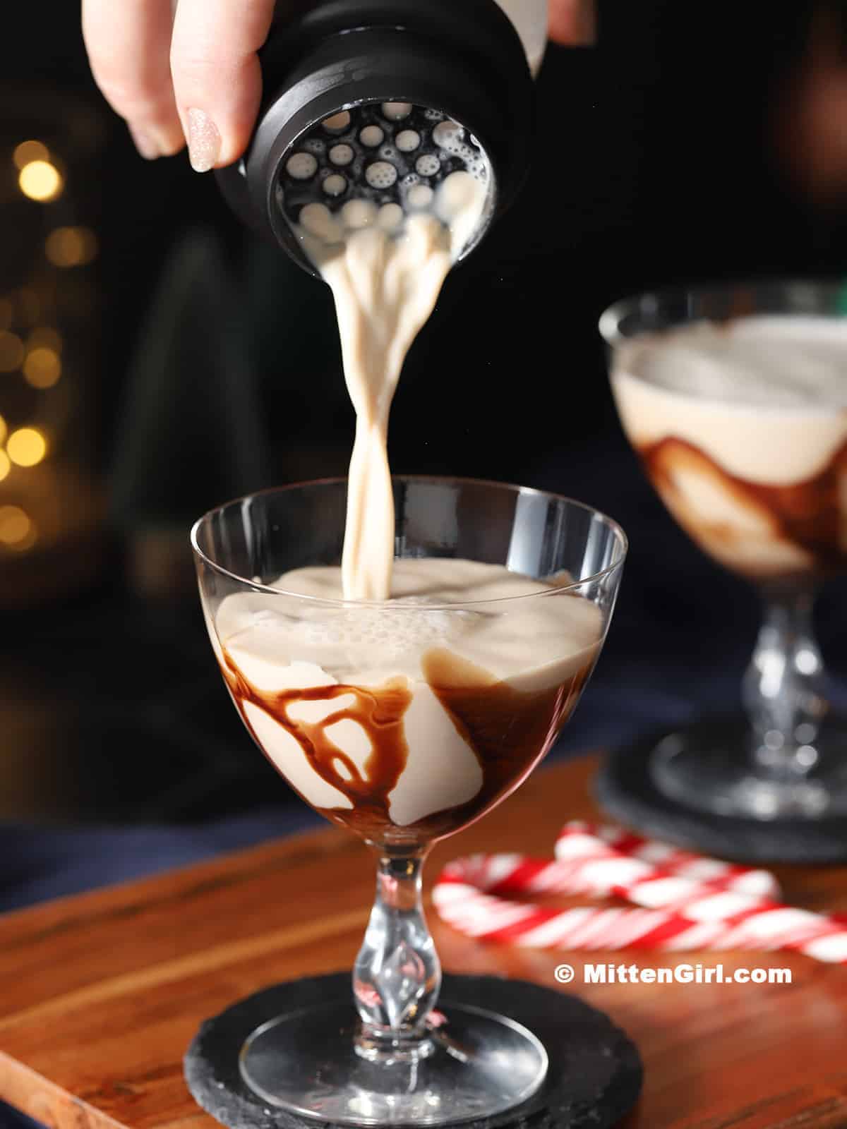 Creamy peppermint mocha martini being poured from a black cocktail shaker into a coupe glass with chocolate syrup drizzle, with candy canes and warm bokeh lights in the background.