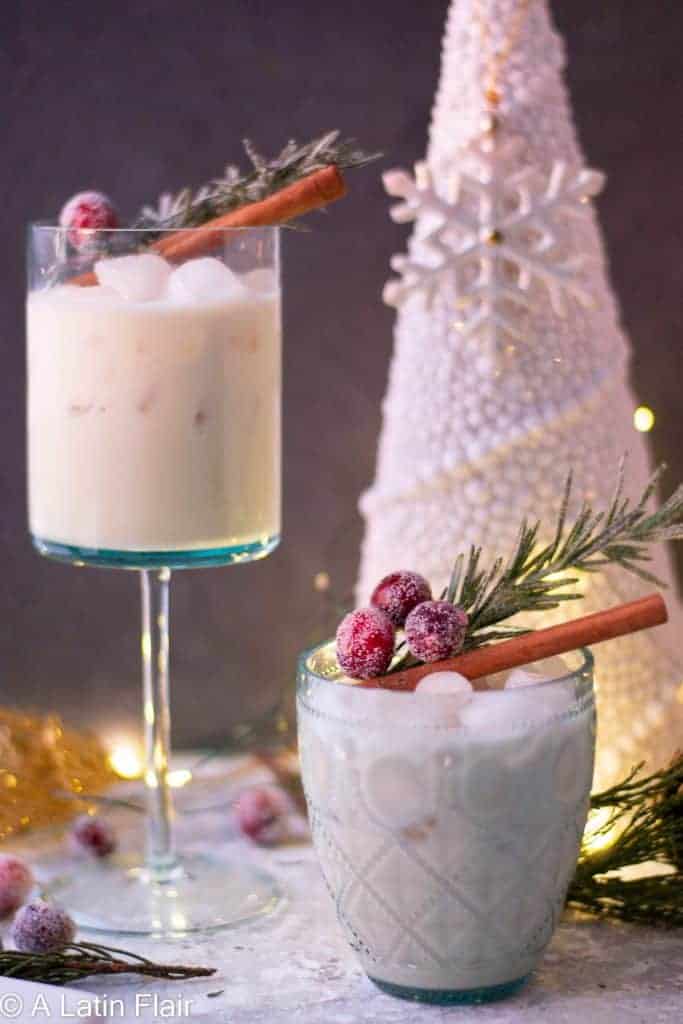 Two glasses of creamy ponche crema (Venezuelan eggnog) garnished with cinnamon sticks, sugared cranberries, and fresh rosemary, displayed with a white Christmas tree and twinkling lights in the background.